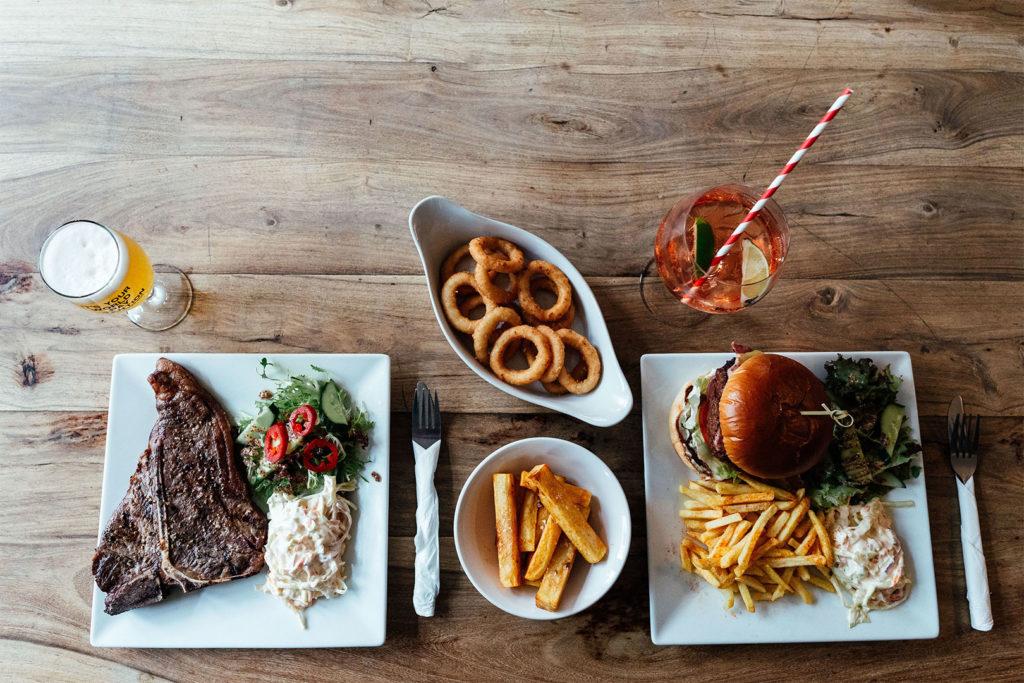 A T bone steak and a burger photographed from above with onion rings and chips