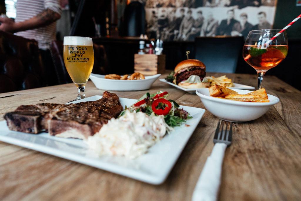 A T bone steak, a burger, onion rings and drinks on a table in the pub