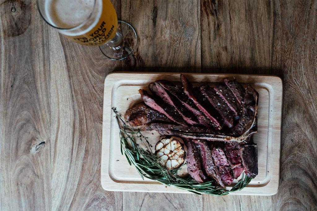 A sliced T bone steak on a wooden board with garlic and herbs